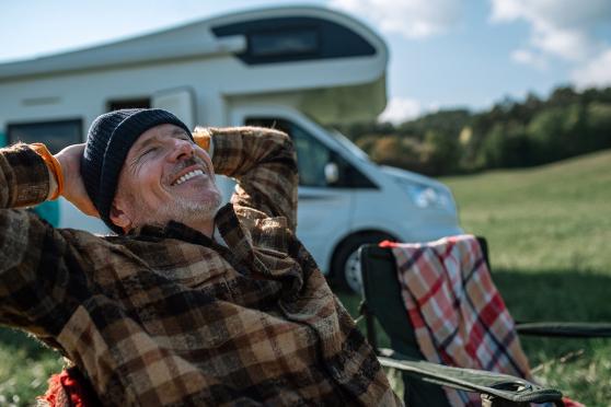 A man sitting in a camping chair, leaned back and smiling, with a trailer in the background